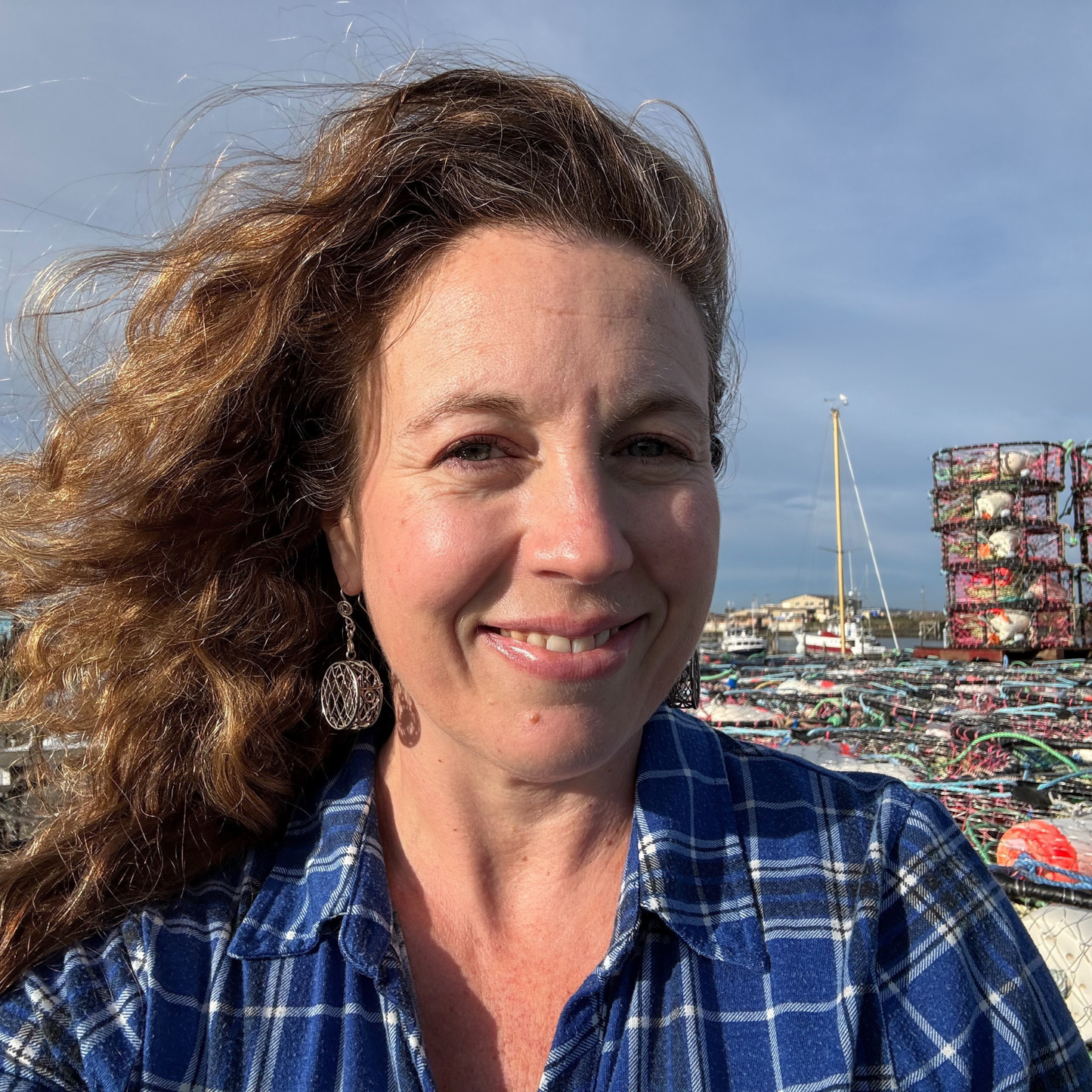 Woman with wavy hair wearing a bright blue plaid shirt, sitting atop a stack of commercial crab pots with the harbor and fishing boats in the background