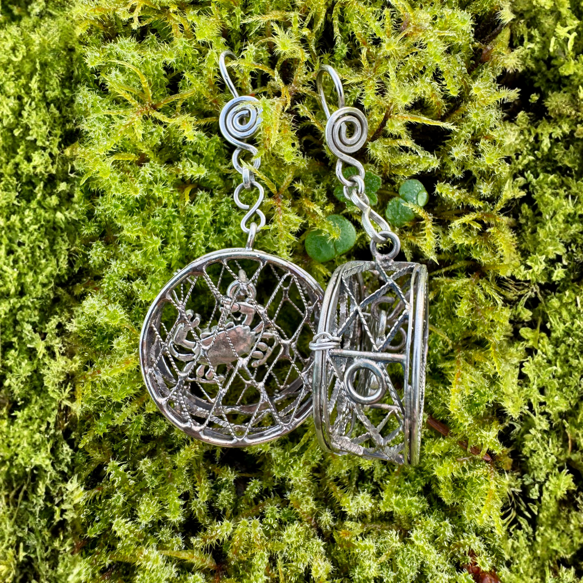 Crab pot earrings with intricate designs on a green moss background one full on view the other is propped on its side to show scale, they are about 1/2 inch wide