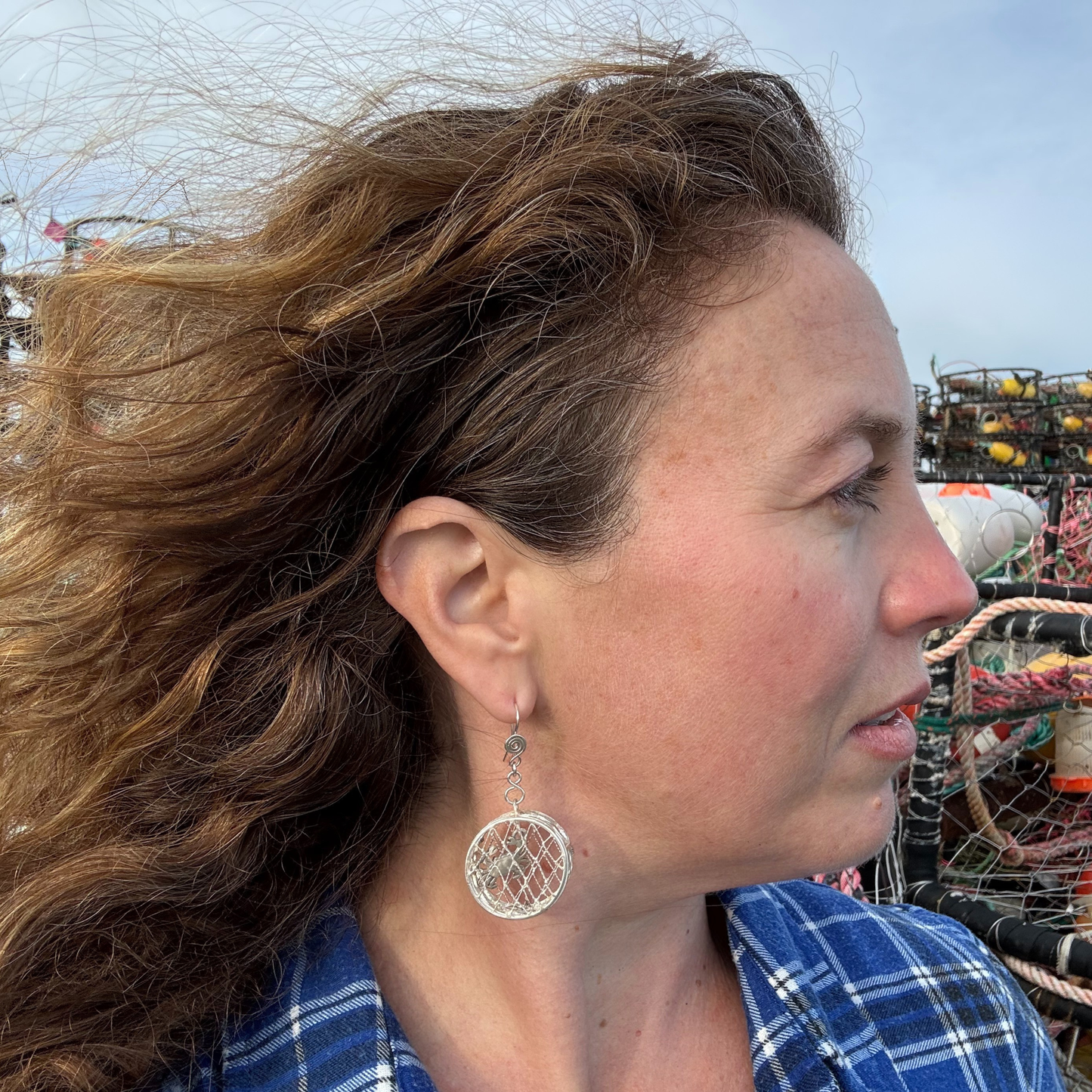 Woman with windblown hair wearing a blue plaid shirt and silver earrings, with a blurred background.